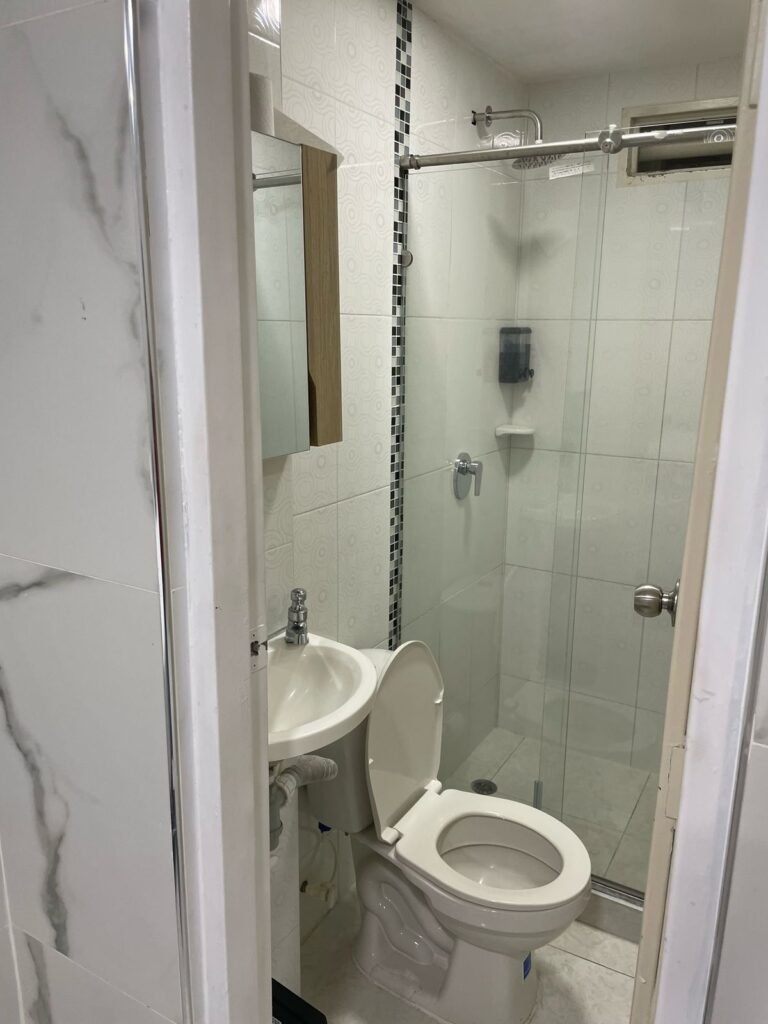 Habitación amoblada de lujo en Cali. Cuenta con una cama amplia, sábanas blancas y toallas dobladas. Las paredes y el piso tienen acabado en mármol con vetas grises. Dispone de un escritorio minimalista con silla ergonómica. La iluminación LED y el ventanal alto garantizan un ambiente moderno y luminoso.