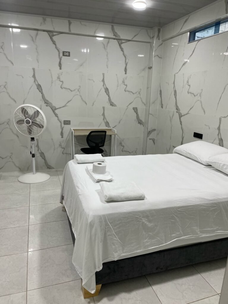Habitación amoblada de lujo en Cali. Cuenta con una cama amplia, sábanas blancas y toallas dobladas. Las paredes y el piso tienen acabado en mármol con vetas grises. Dispone de un escritorio minimalista con silla ergonómica. La iluminación LED y el ventanal alto garantizan un ambiente moderno y luminoso.