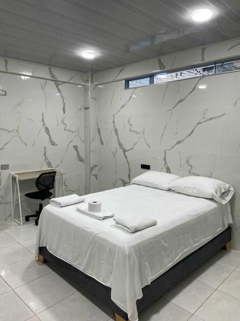 Habitación amoblada de lujo en Cali. Cuenta con una cama amplia, sábanas blancas y toallas dobladas. Las paredes y el piso tienen acabado en mármol con vetas grises. Dispone de un escritorio minimalista con silla ergonómica. La iluminación LED y el ventanal alto garantizan un ambiente moderno y luminoso.