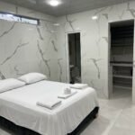 Habitación amoblada de lujo en Cali. Cuenta con una cama amplia, sábanas blancas y toallas dobladas. Las paredes y el piso tienen acabado en mármol con vetas grises. Dispone de un escritorio minimalista con silla ergonómica. La iluminación LED y el ventanal alto garantizan un ambiente moderno y luminoso.