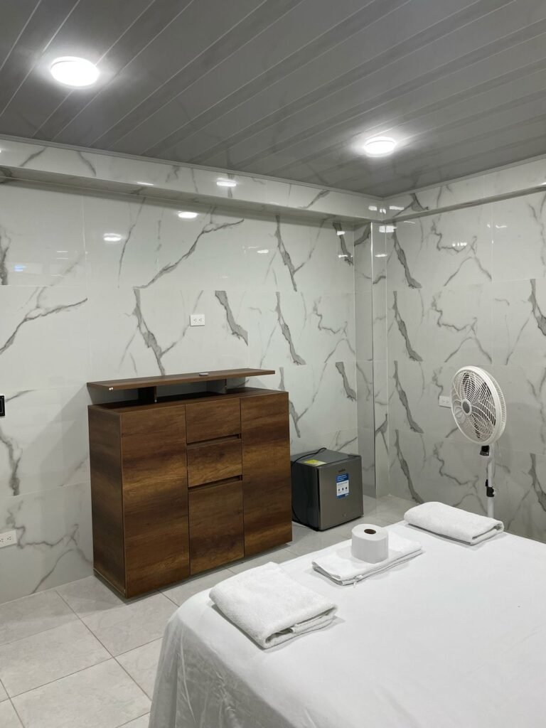 Habitación amoblada de lujo en Cali. Cuenta con una cama amplia, sábanas blancas y toallas dobladas. Las paredes y el piso tienen acabado en mármol con vetas grises. Dispone de un escritorio minimalista con silla ergonómica. La iluminación LED y el ventanal alto garantizan un ambiente moderno y luminoso.